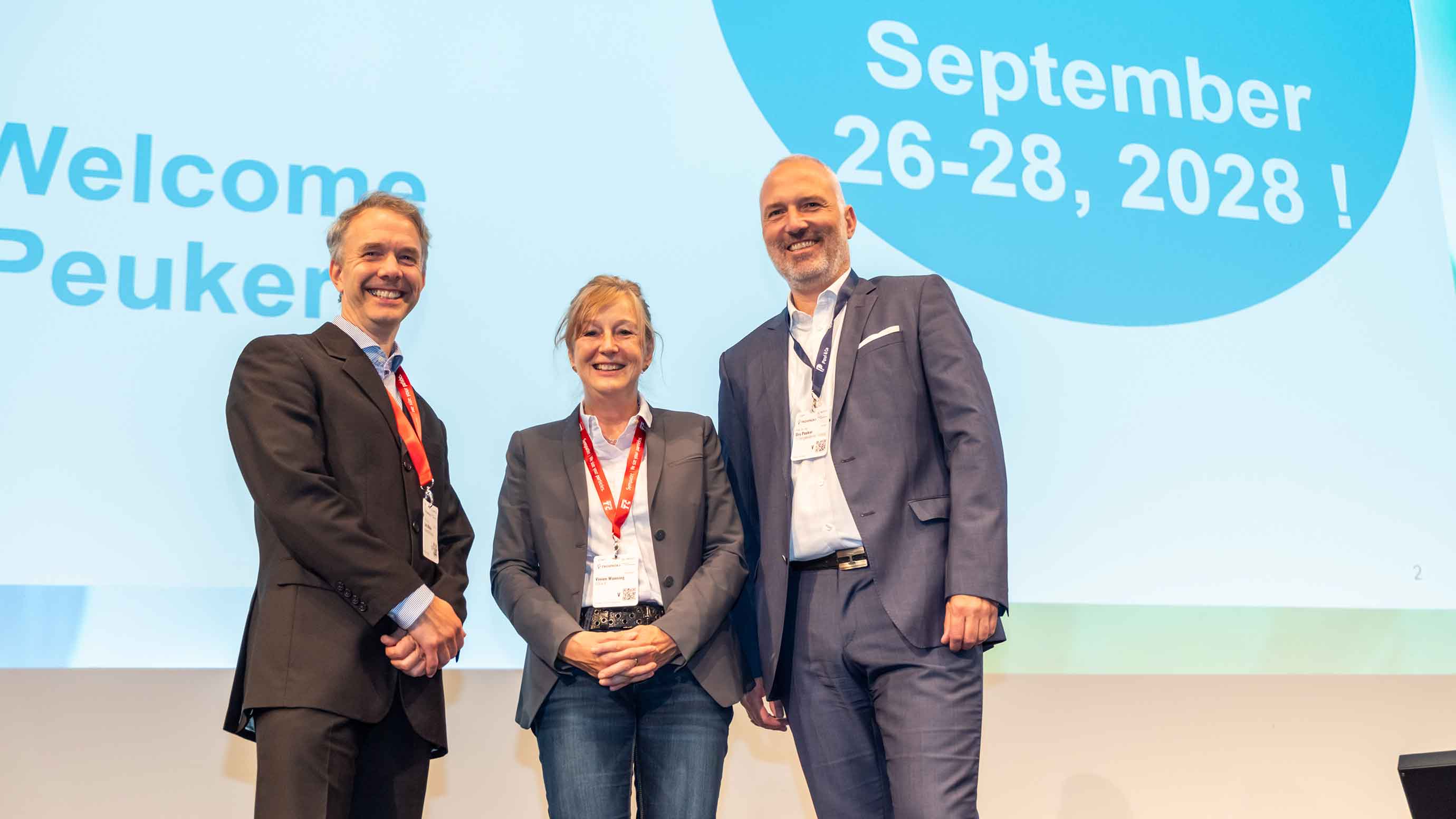 Three people standing on a conference stage in front of a large presentation screen. All three individuals wear business attire and event lanyards.