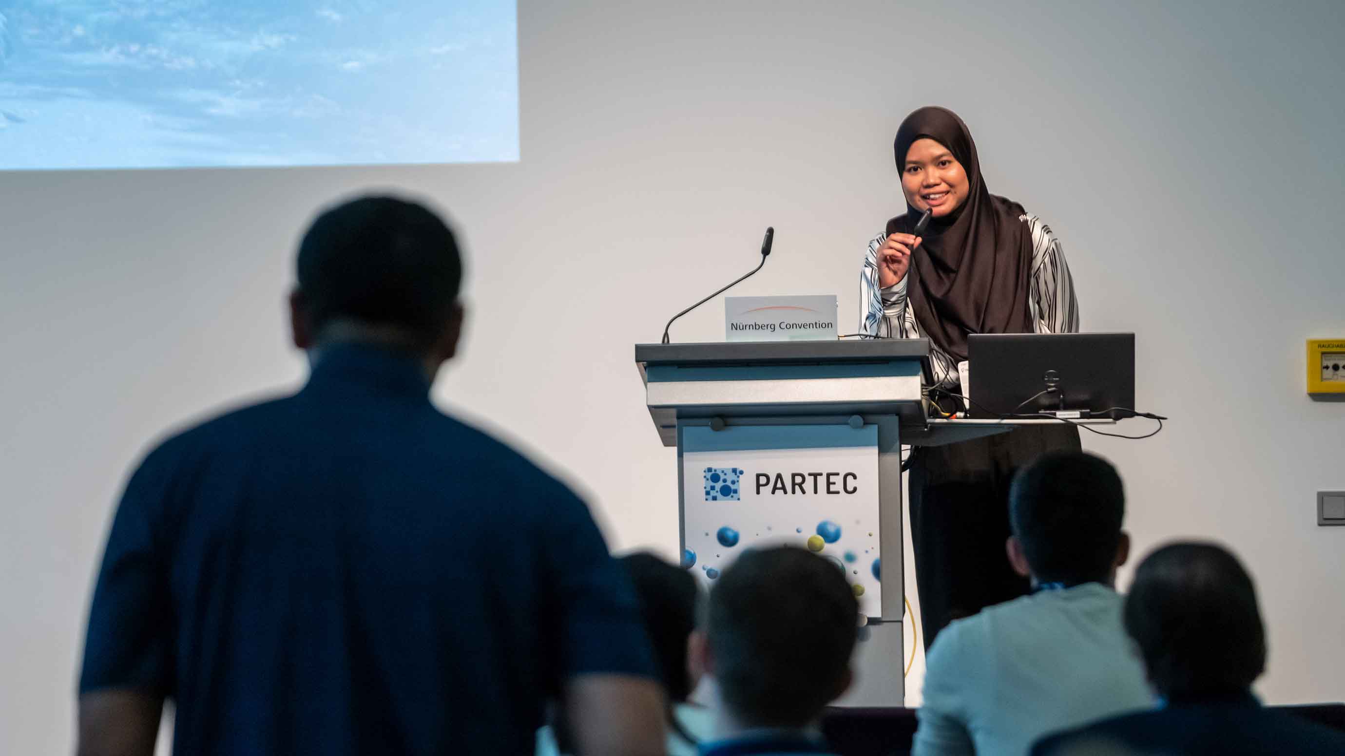 A speaker standing at a podium labeled “PARTEC” presents to an audience in a conference room. A microphone, laptop, and nameplate reading “Nuremberg Convention” are visible on the podium. Audience members are seated in the foreground, facing the stage.