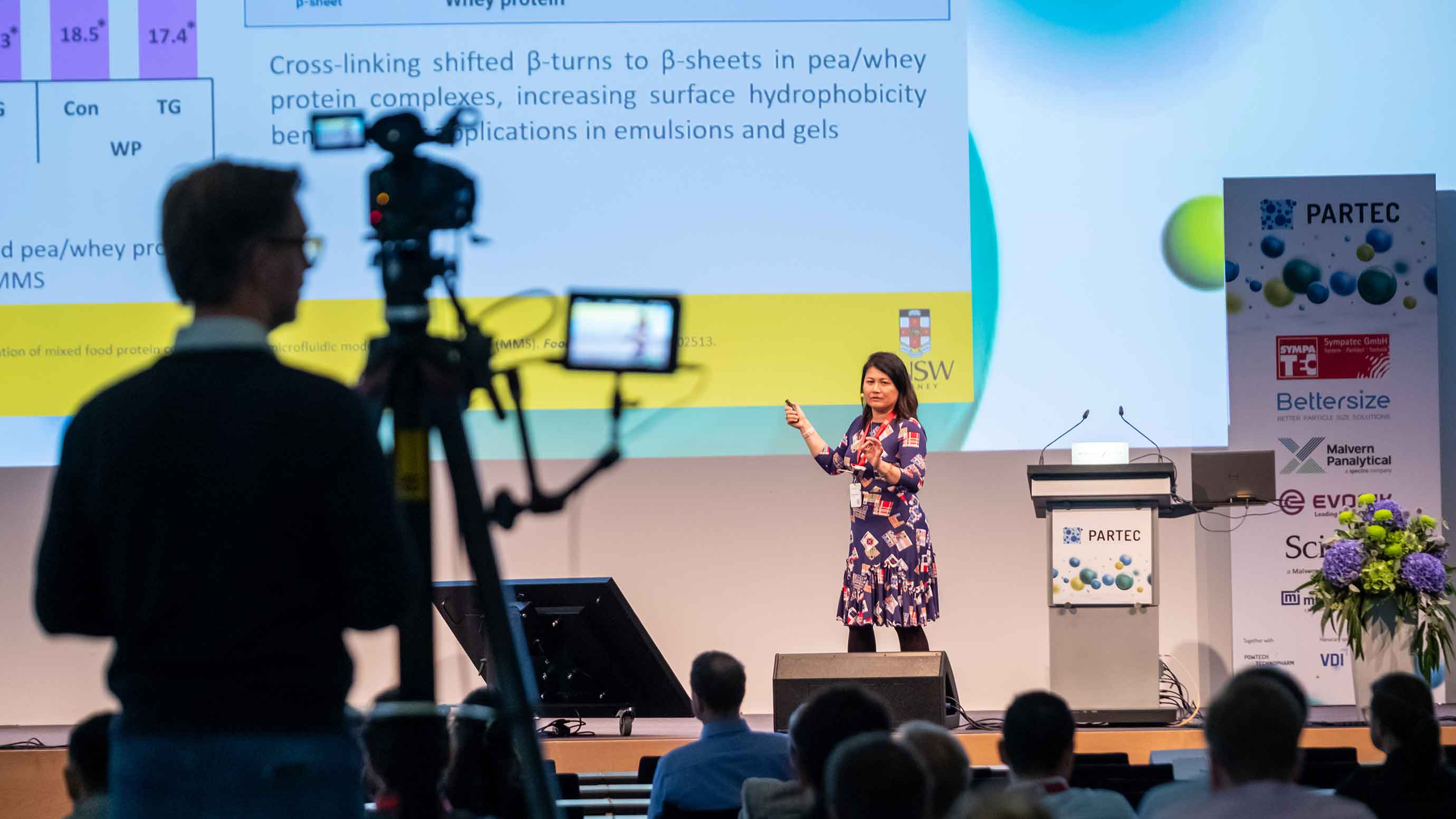 A person stands on a conference stage giving a presentation, pointing toward a large projected slide containing scientific text and data. Audience members are seated in the foreground, and a camera operator films the session. A podium labeled “PARTEC” and a display stand with various sponsor logos are visible on the right.