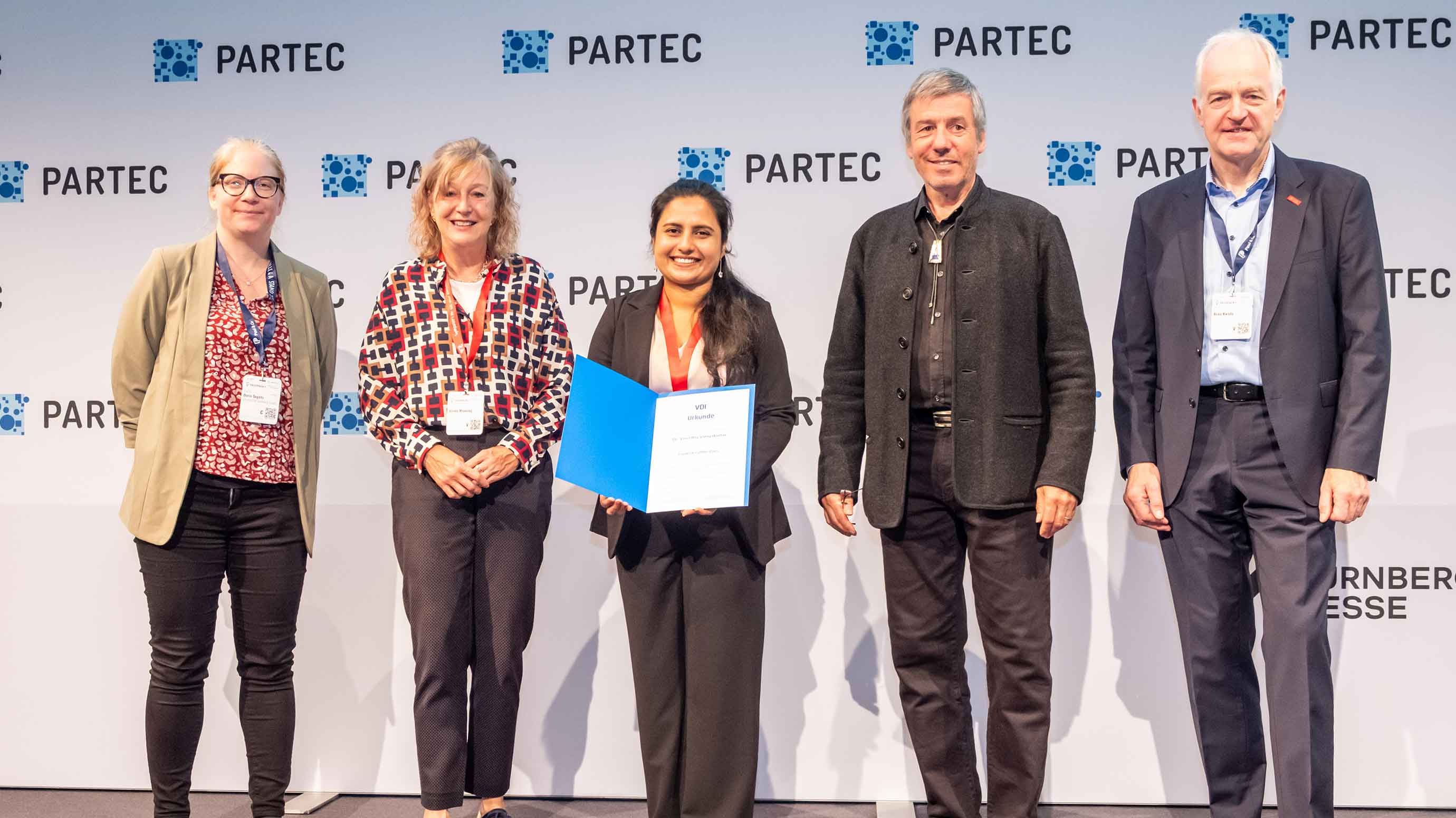 A group of five people standing on a stage in front of a backdrop displaying multiple “PARTEC” logos. The person in the center holds an open certificate folder. All individuals wear event badges, and the scene appears to depict an award or recognition moment.